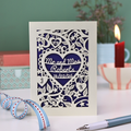A personalised paper cut wedding card with floral heart design, sitting on a white desk with ribbons, stationery items and plant on a green background. Card is cream with "Mr and Mrs Roberts, 14/02/26" on 3 lines of text inside a heart shape with intricate floral cut out border.