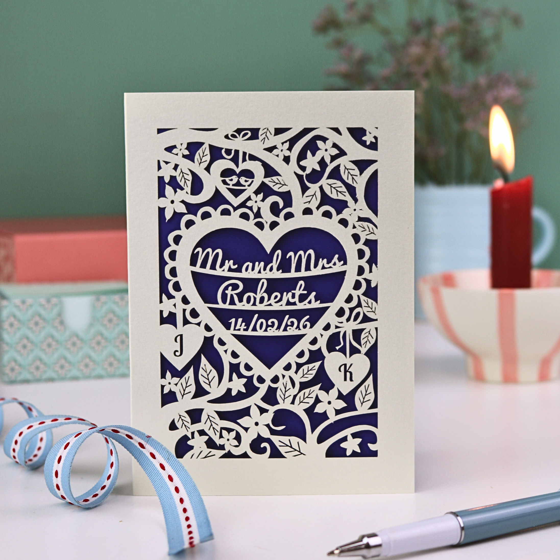 A personalised paper cut wedding card with floral heart design, sitting on a white desk with ribbons, stationery items and plant on a green background. Card is cream with "Mr and Mrs Roberts, 14/02/26" on 3 lines of text inside a heart shape with intricate floral cut out border.