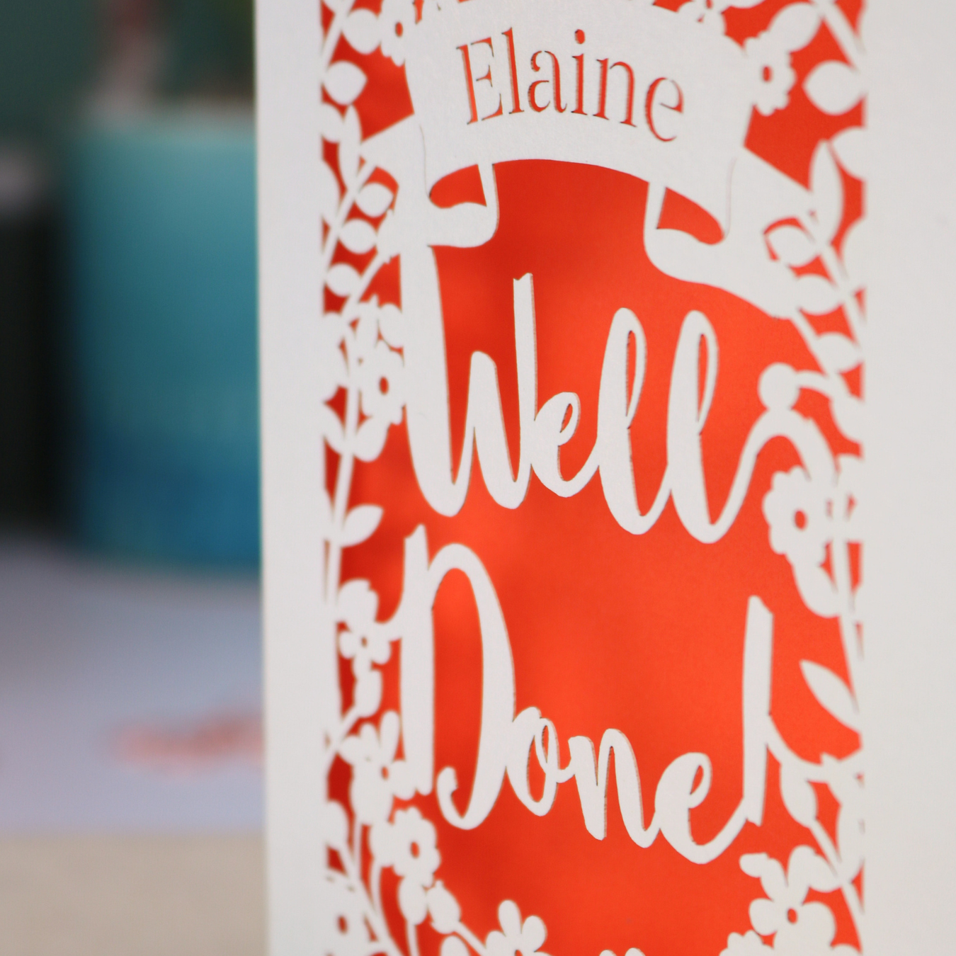 Laser cut Greeting card with 'Well Done' and 'Elaine' text on a desk with stationery items.