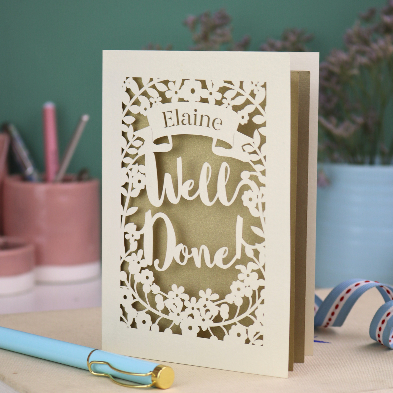Laser cut Greeting card with 'Well Done' and 'Elaine' text on a desk with stationery items.