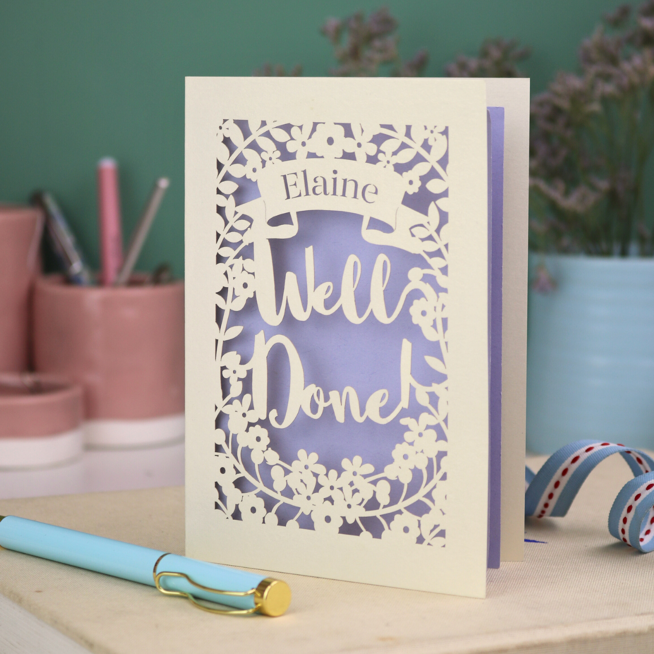 Laser cut Greeting card with 'Well Done' and 'Elaine' text on a desk with stationery items.