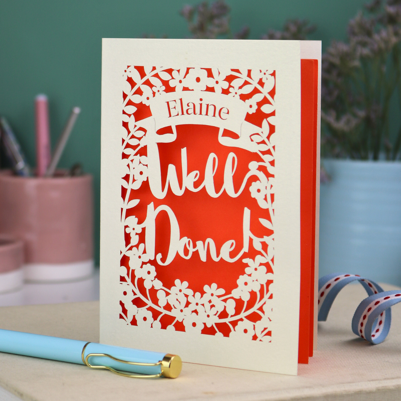 Laser cut Greeting card with 'Well Done' and 'Elaine' text on a desk with stationery items.