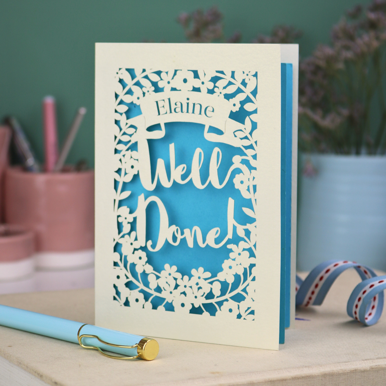 Laser cut Greeting card with 'Well Done' and 'Elaine' text on a desk with stationery items.
