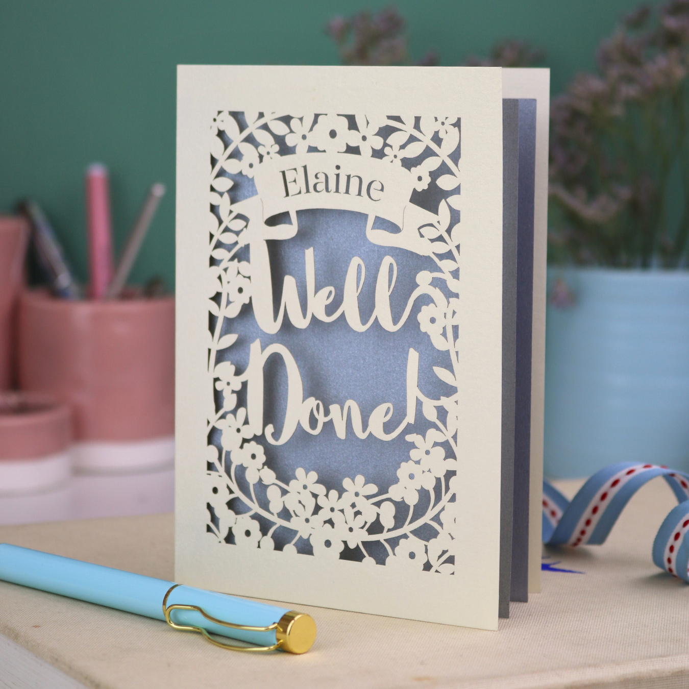 Laser cut Greeting card with 'Well Done' and 'Elaine' text on a desk with stationery items.