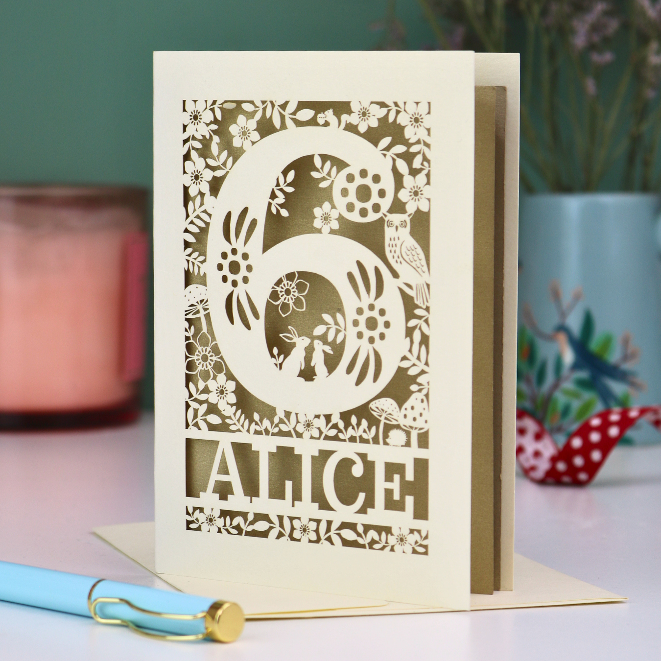 Laser cut 6th Birthday card with decorative number 6 and name 'Alice', and woodland design, on a table with a pen and plant in the background.