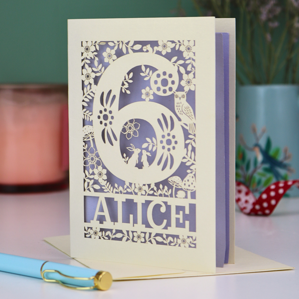 Laser cut 6th Birthday card with decorative number 6 and name 'Alice', and woodland design, on a table with a pen and plant in the background.