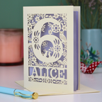 Laser cut 6th Birthday card with decorative number 6 and name 'Alice', and woodland design, on a table with a pen and plant in the background.