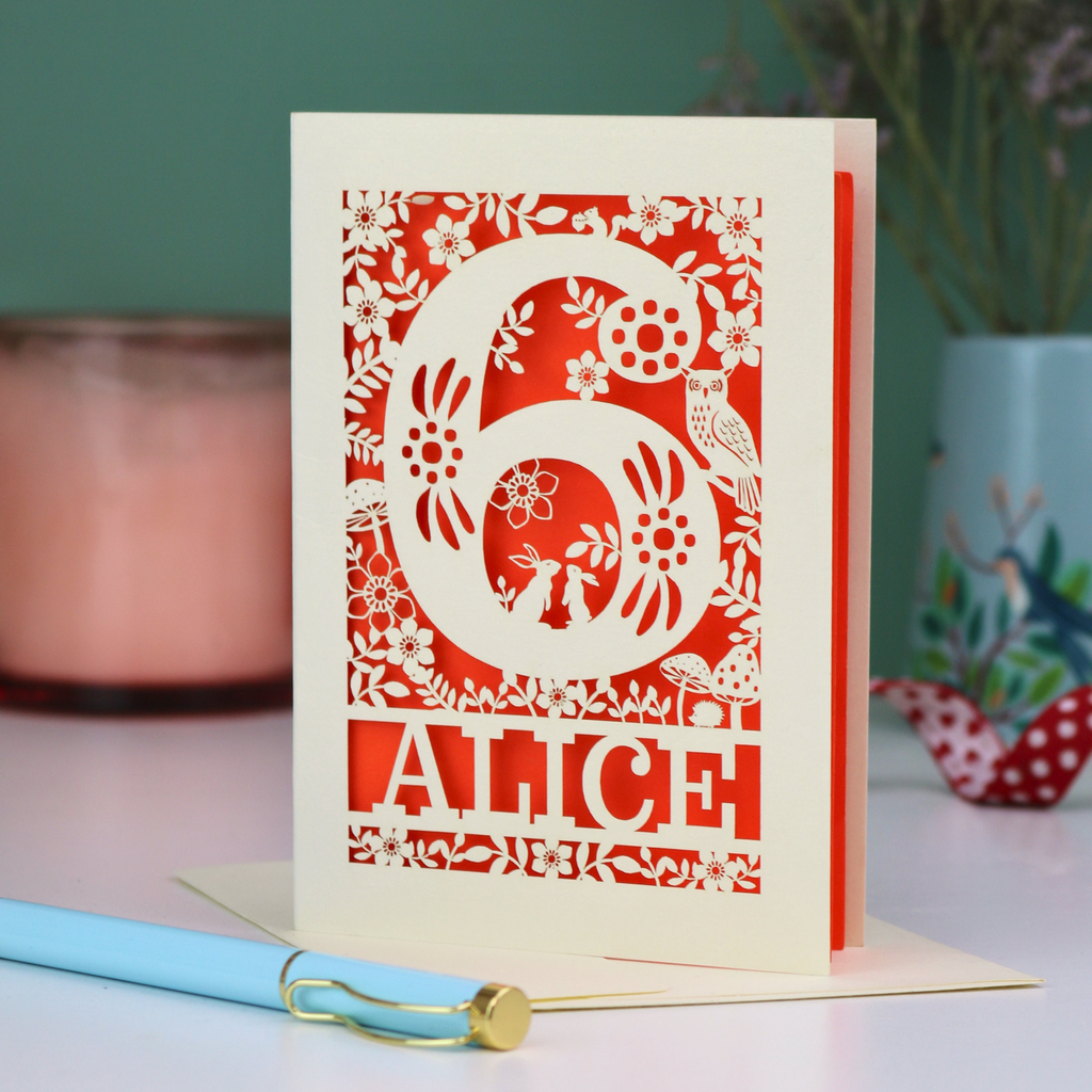 Laser cut 6th Birthday card with decorative number 6 and name 'Alice', and woodland design, on a table with a pen and plant in the background.