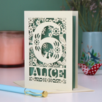 Laser cut 6th Birthday card with decorative number 6 and name 'Alice', and woodland design, on a table with a pen and plant in the background.