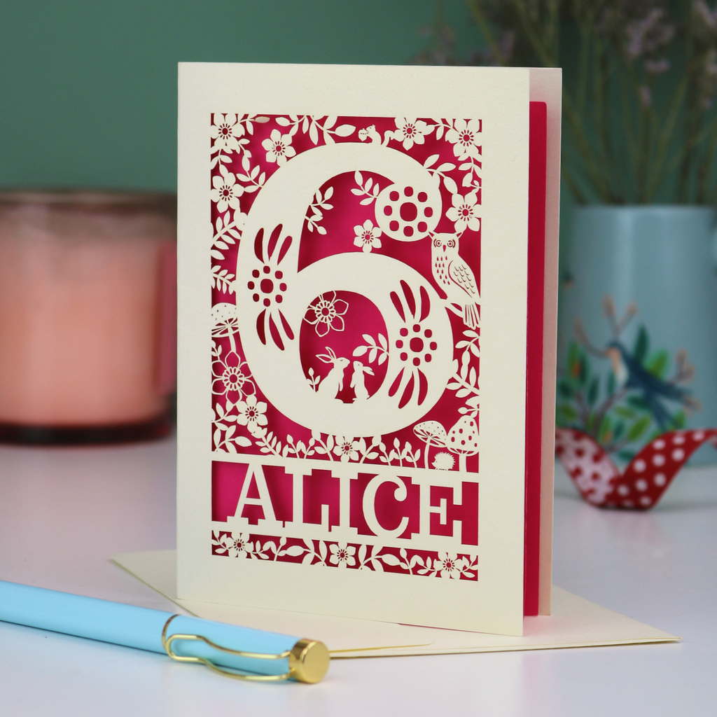 Laser cut 6th Birthday card with decorative number 6 and name 'Alice', and woodland design, on a table with a pen and plant in the background.