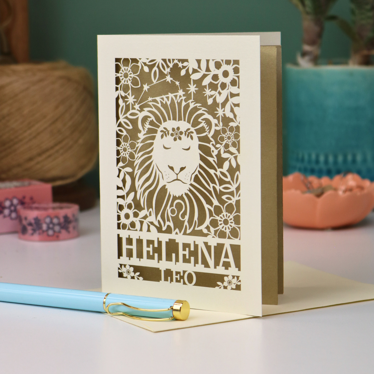 A cream Leo birthday card with a red laser-cut lion and floral design, standing upright on a table with a light blue pen in the foreground and soft-focus decorative objects in the background.