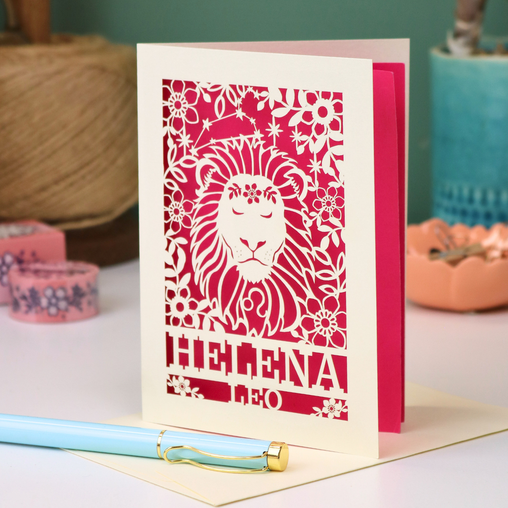 A cream Leo birthday card with a red laser-cut lion and floral design, standing upright on a table with a light blue pen in the foreground and soft-focus decorative objects in the background.