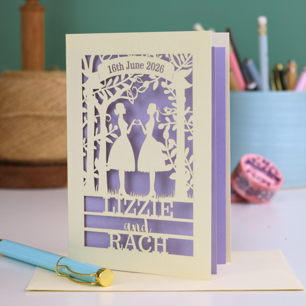 Personalized lesbian wedding card with cut-out design of two people and text, placed on a surface with stationery items.