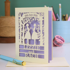 Personalized lesbian wedding card with cut-out design of two people and text, placed on a surface with stationery items.