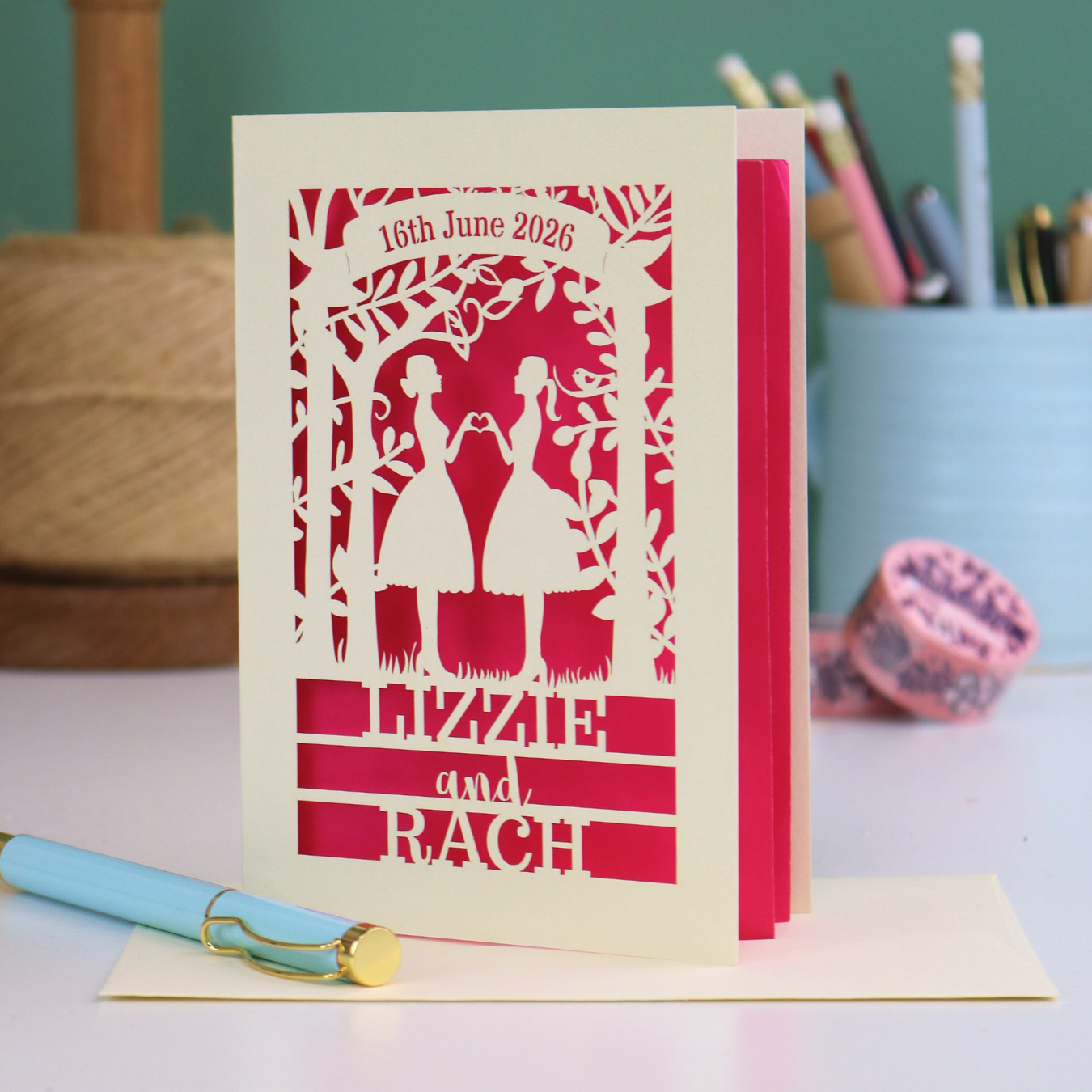 Personalised card with cut-out design of two women holding hands, text '16th June 2026', 'Lizzie and Rach', on a pink background.