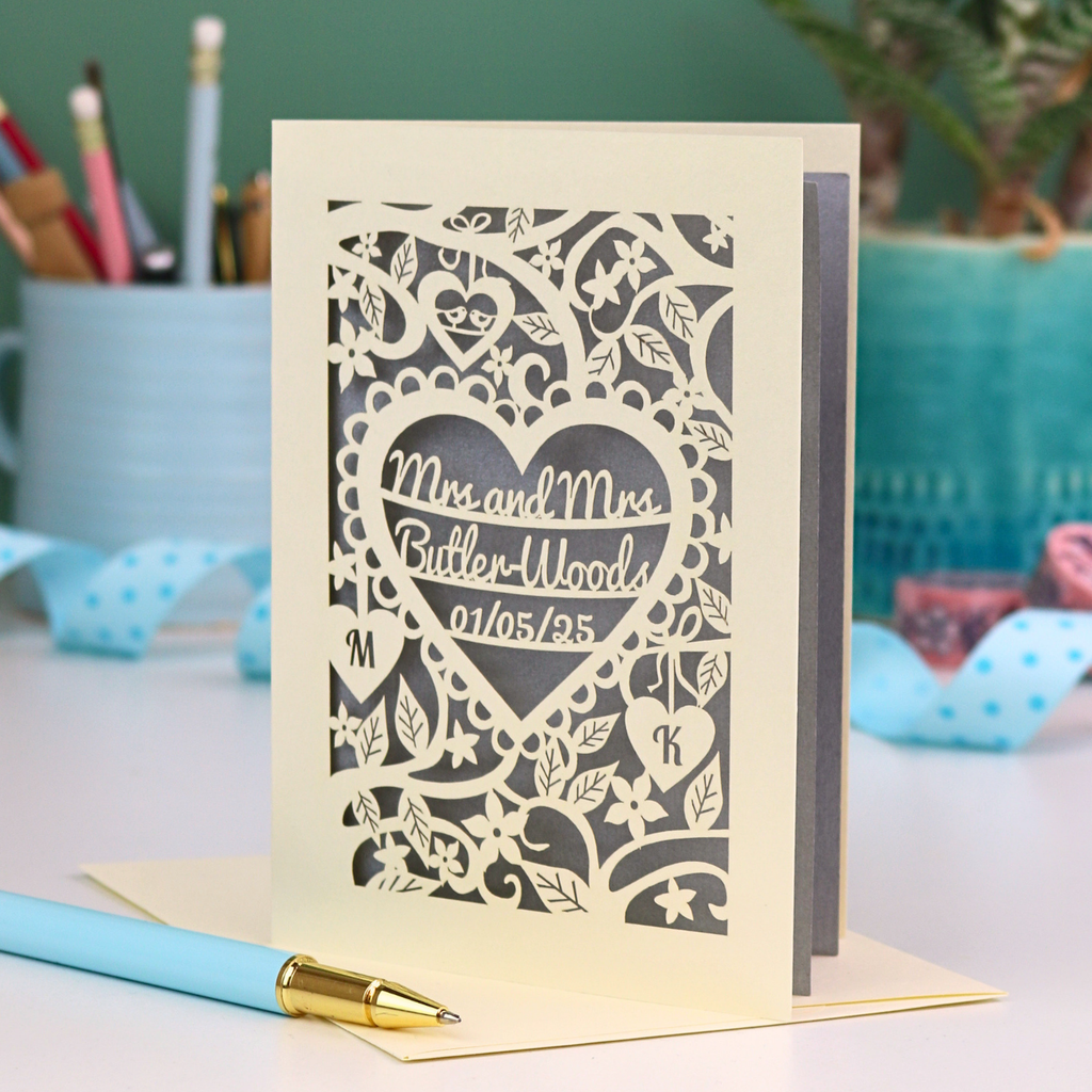 Laser-cut Mrs and Mrs anniversary card with personalized text, 'Mrs and Mrs Butler-Woods, 01/05/25', surrounded by leaves and flowers,  on a desk with stationery items.