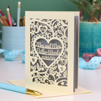 Laser-cut Mrs and Mrs anniversary card with personalized text, 'Mrs and Mrs Butler-Woods, 01/05/25', surrounded by leaves and flowers,  on a desk with stationery items.