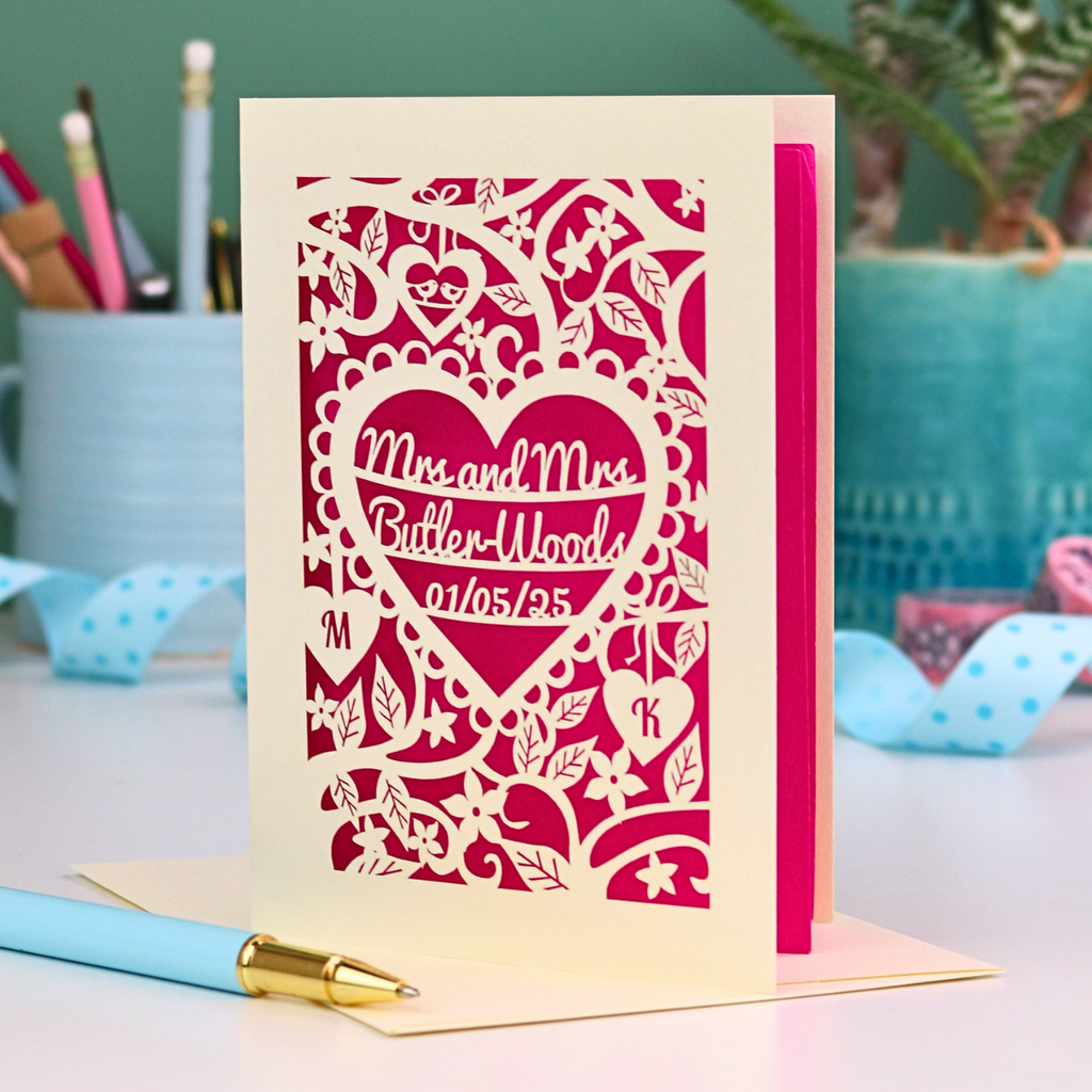 Laser-cut lesbian anniversary card with personalized text, 'Mrs and Mrs Butler-Woods, 01/05/25', surrounded by leaves and flowers,  on a desk with stationery items.