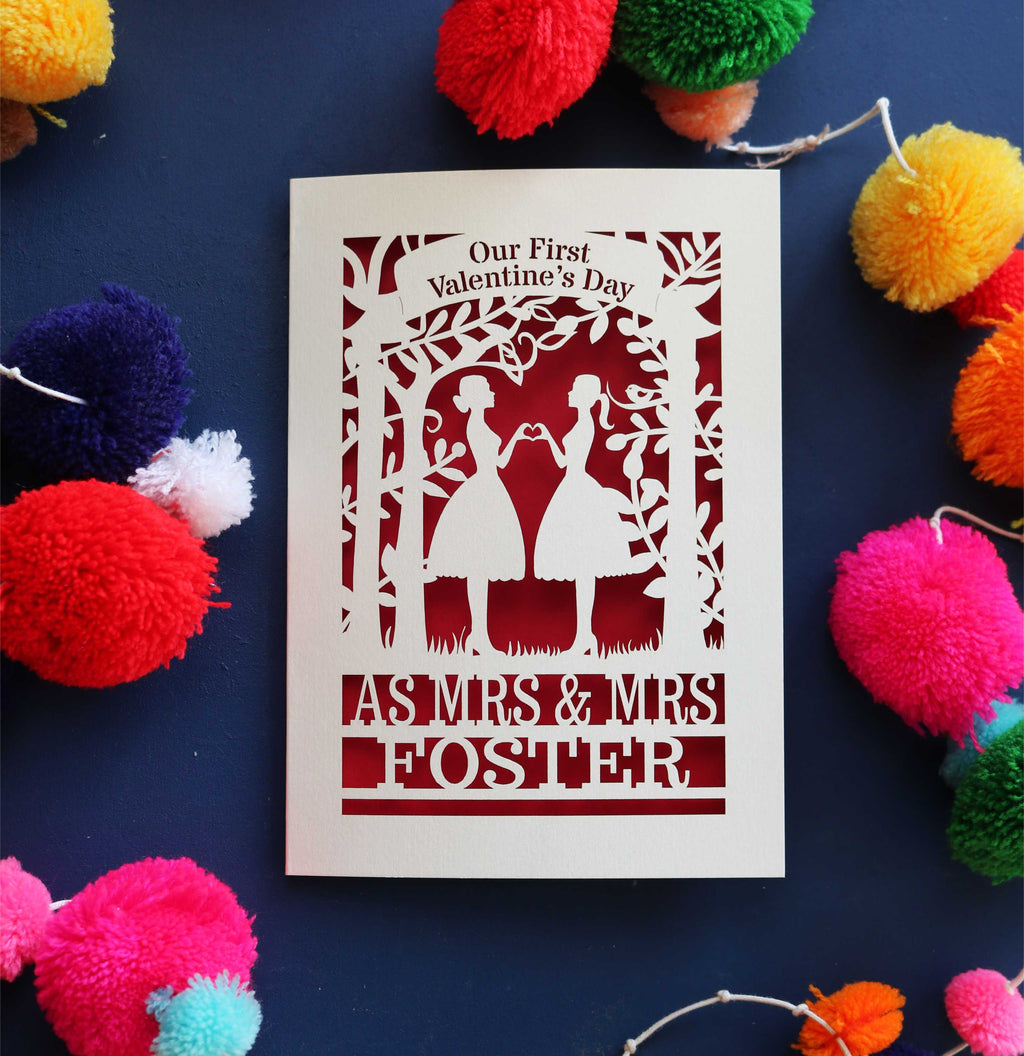 A personalised Valentine's day card with a laser cut design of two women making a heart shape, with a blank space for a personal message, and brightly coloured pom-pom decorations around it.