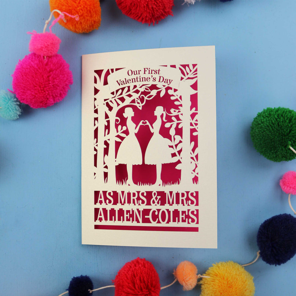 A personalised lesbian first Valentine's day card with a laser cut design of two women making a heart shape, with a blank space for a personal message, and brightly coloured pom-pom decorations around it.