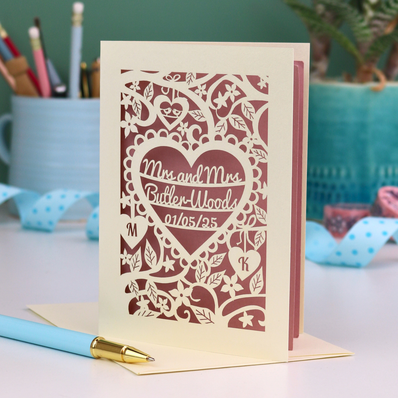 Laser-cut Mrs and Mrs anniversary card with personalized text, 'Mrs and Mrs Butler-Woods, 01/05/25', surrounded by leaves and flowers,  on a desk with stationery items.