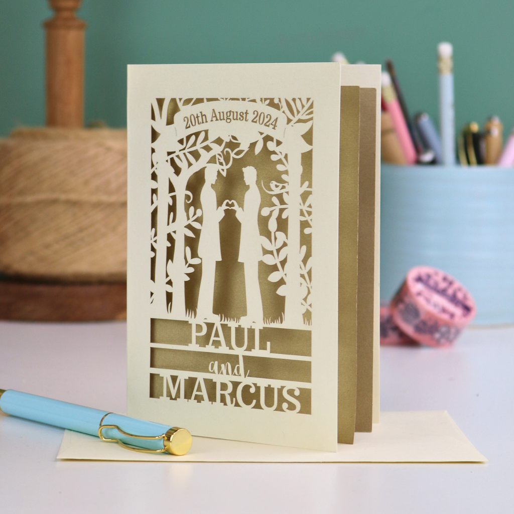 Personalized card with laser-cut design of two male figures holding hands, with text '20th August 2024', 'PAUL', and 'MARCUS' on a desk.