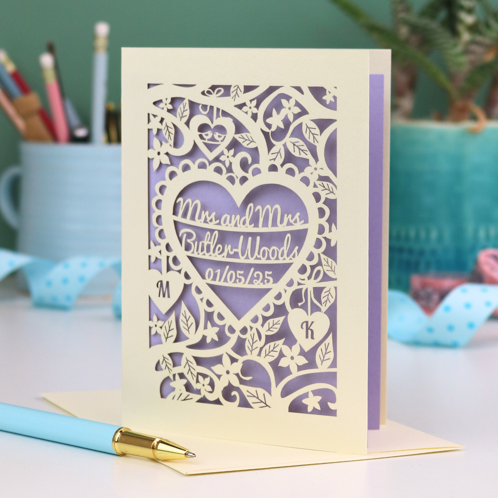 Laser-cut Mrs and Mrs LGBTQIA+ anniversary card with personalized text, 'Mrs and Mrs Butler-Woods, 01/05/25', surrounded by leaves and flowers,  on a desk with stationery items.
