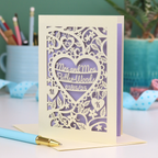 Laser-cut Mrs and Mrs LGBTQIA+ anniversary card with personalized text, 'Mrs and Mrs Butler-Woods, 01/05/25', surrounded by leaves and flowers,  on a desk with stationery items.