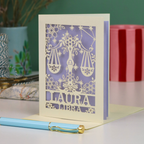 A personalized laser-cut Libra birthday card with a Libra zodiac design, featuring a lavender purple background and a silhouette of the Libra scales, with the name 'LAURA' and text 'LIBRA' cut out at the bottom. A light blue pen with a gold clip rests in front of the card on a white surface, with a green wall and decorative background elements visible.