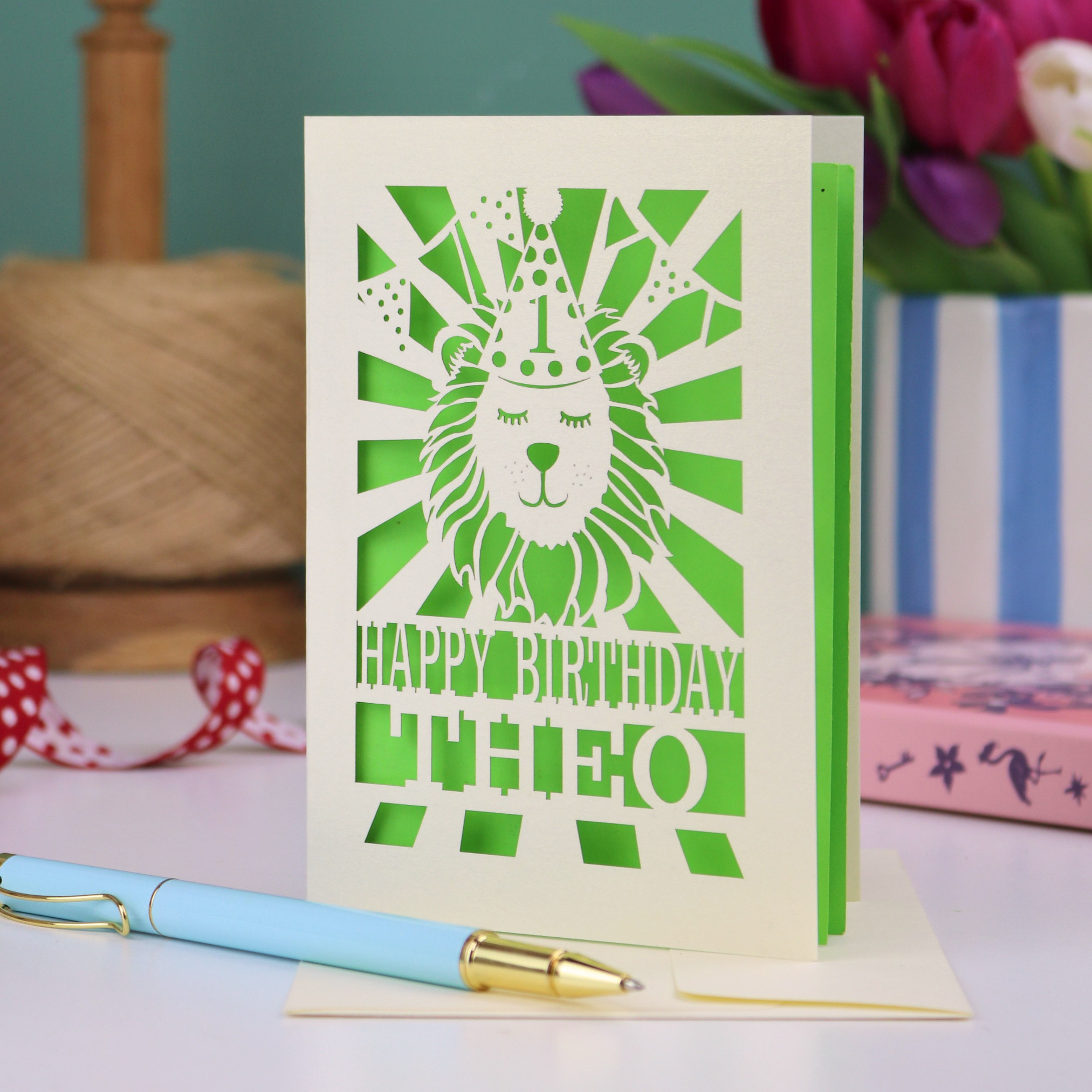 A personalised laser cut first birthday card for boys, with cut out design of a lion in a party hat, and text "Happy Birthday Theo"