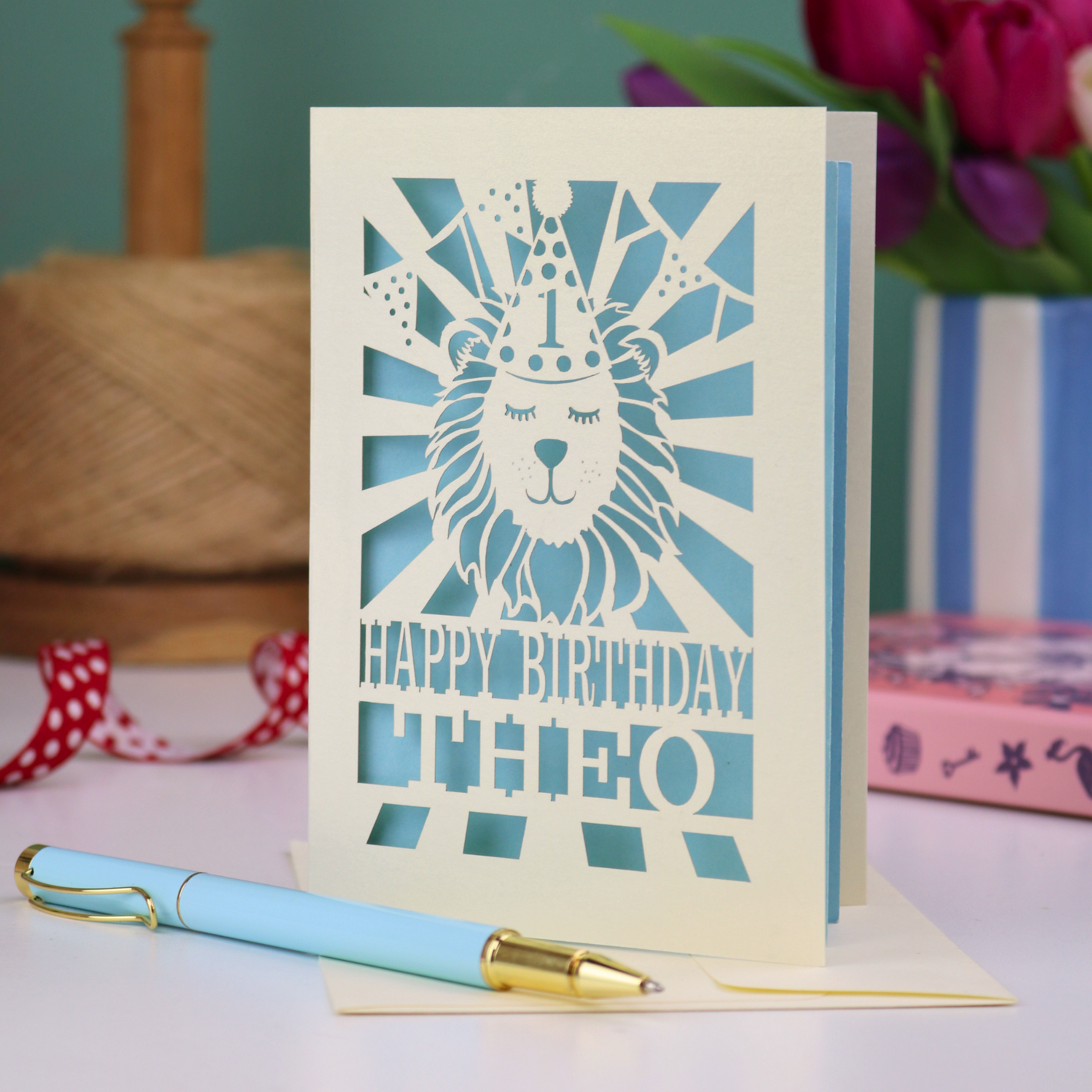 A personalised laser cut 1st birthday card with cut out design of a lion in a party hat, and text "Happy Birthday Theo"
