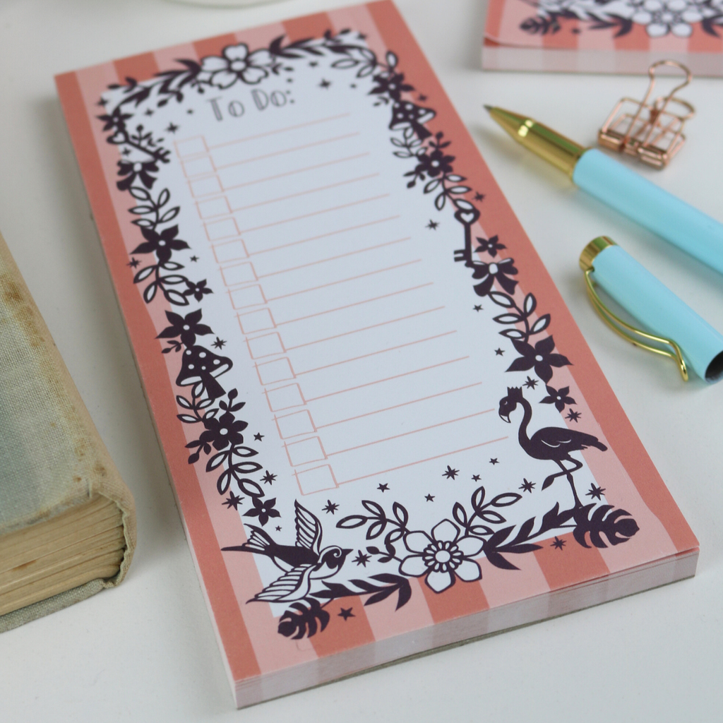 A close up detail of a notepad with pink striped border and purple floral illustrations, with "To Do:" at the top and lines with check boxes