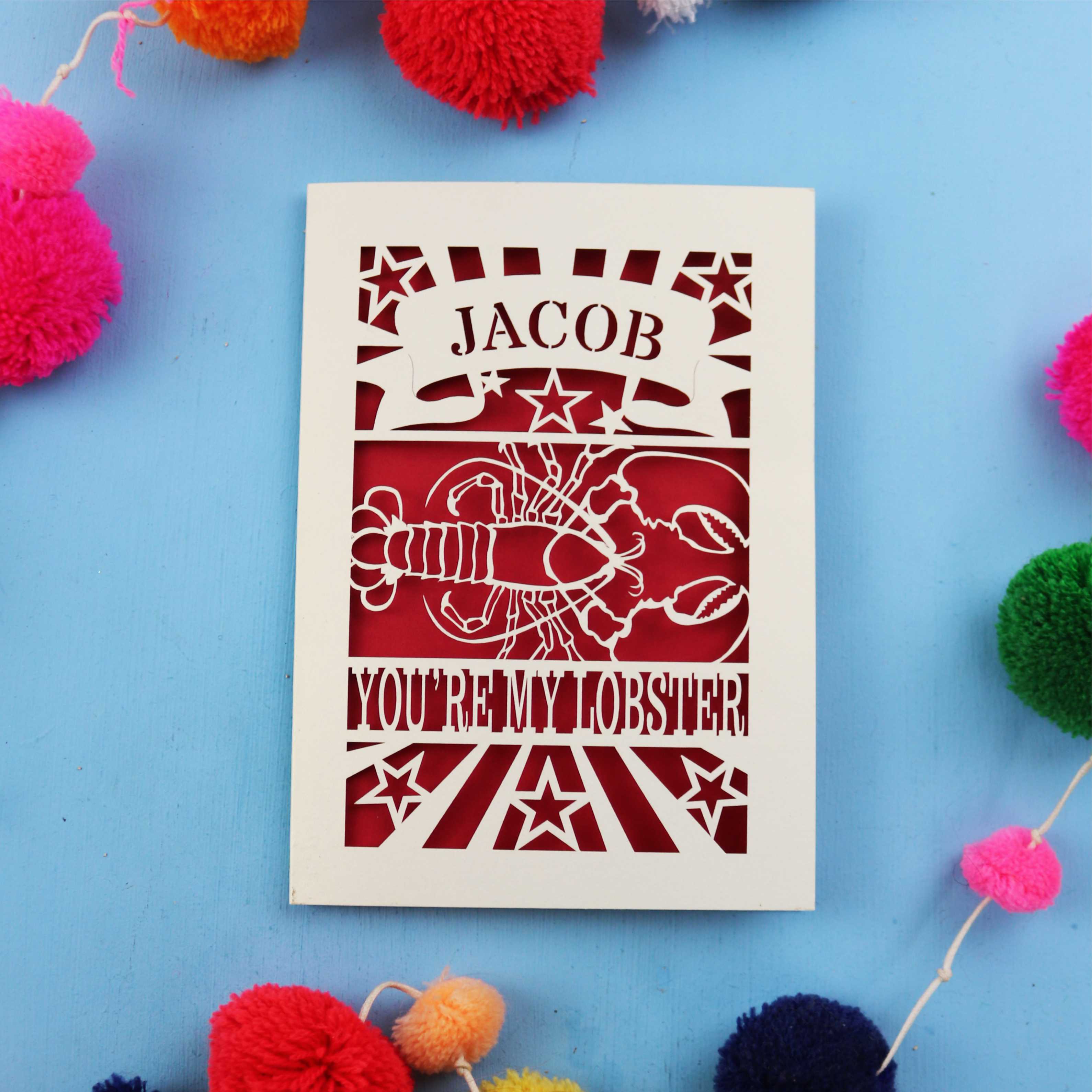 Laser Cut Valentine's Card with 'Jacob, you're my lobster' design on a blue background with colourful pom-poms.