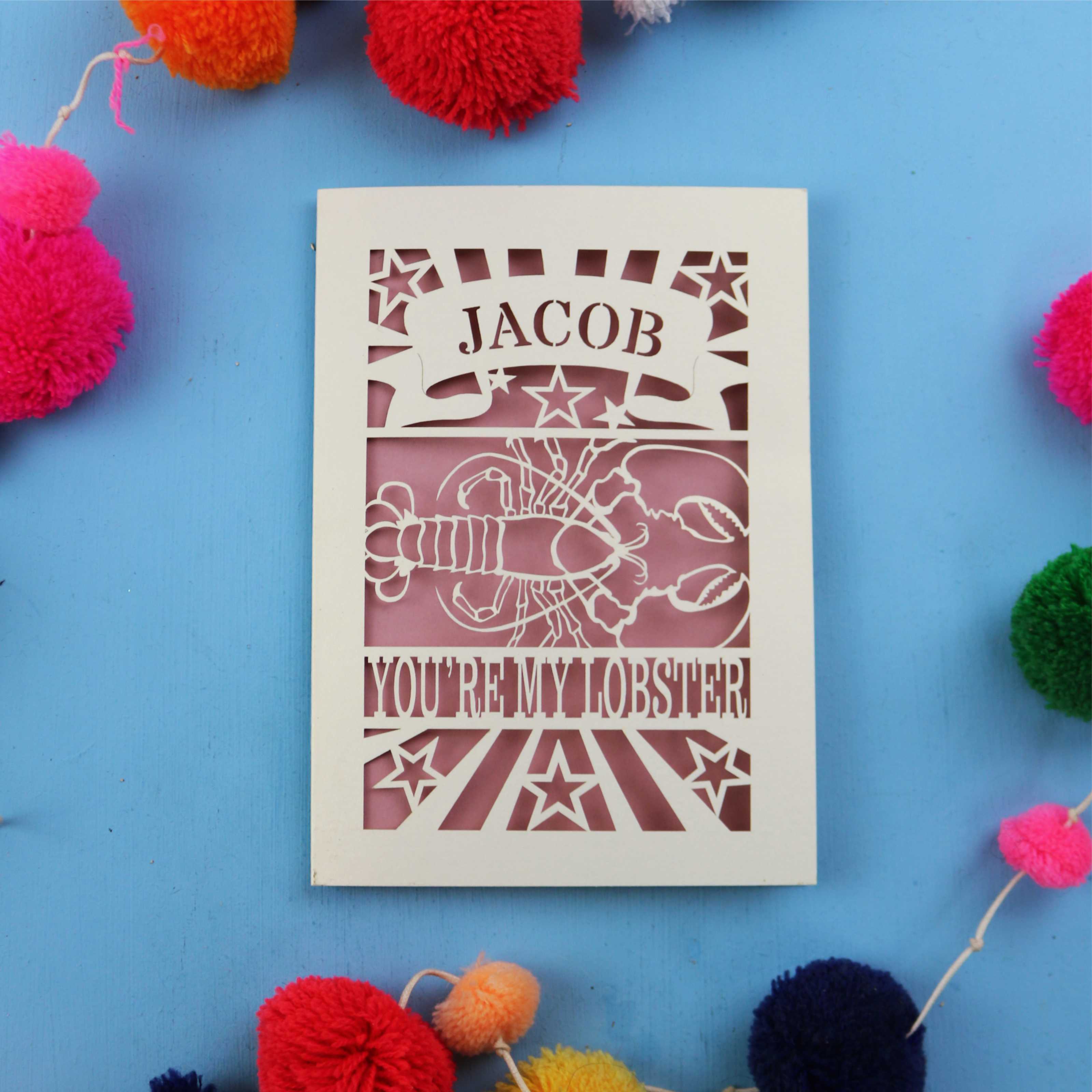 Paper Cut Valentine's Card with 'Jacob, you're my lobster' design on a blue background with colourful pom-poms.