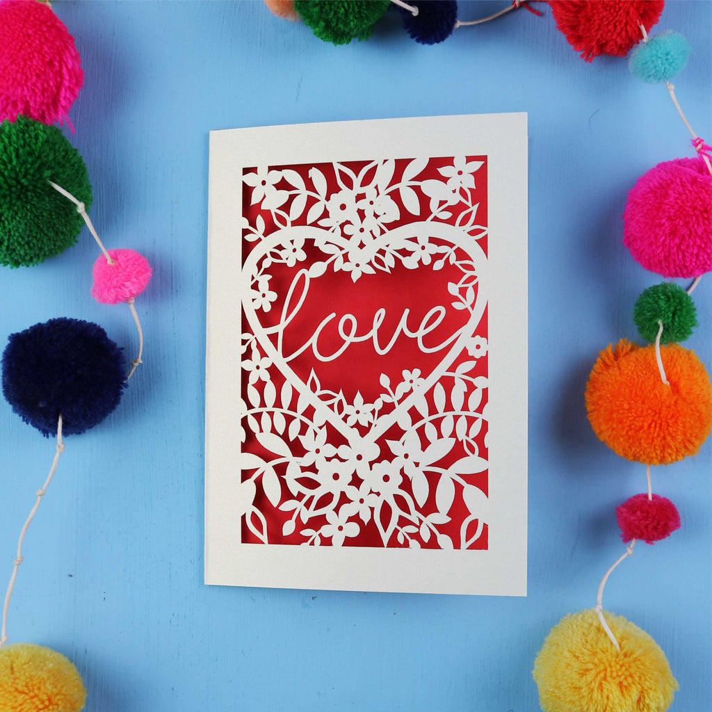 Laser cut Card with 'love' design surrounded by colourful pom-poms on a blue background