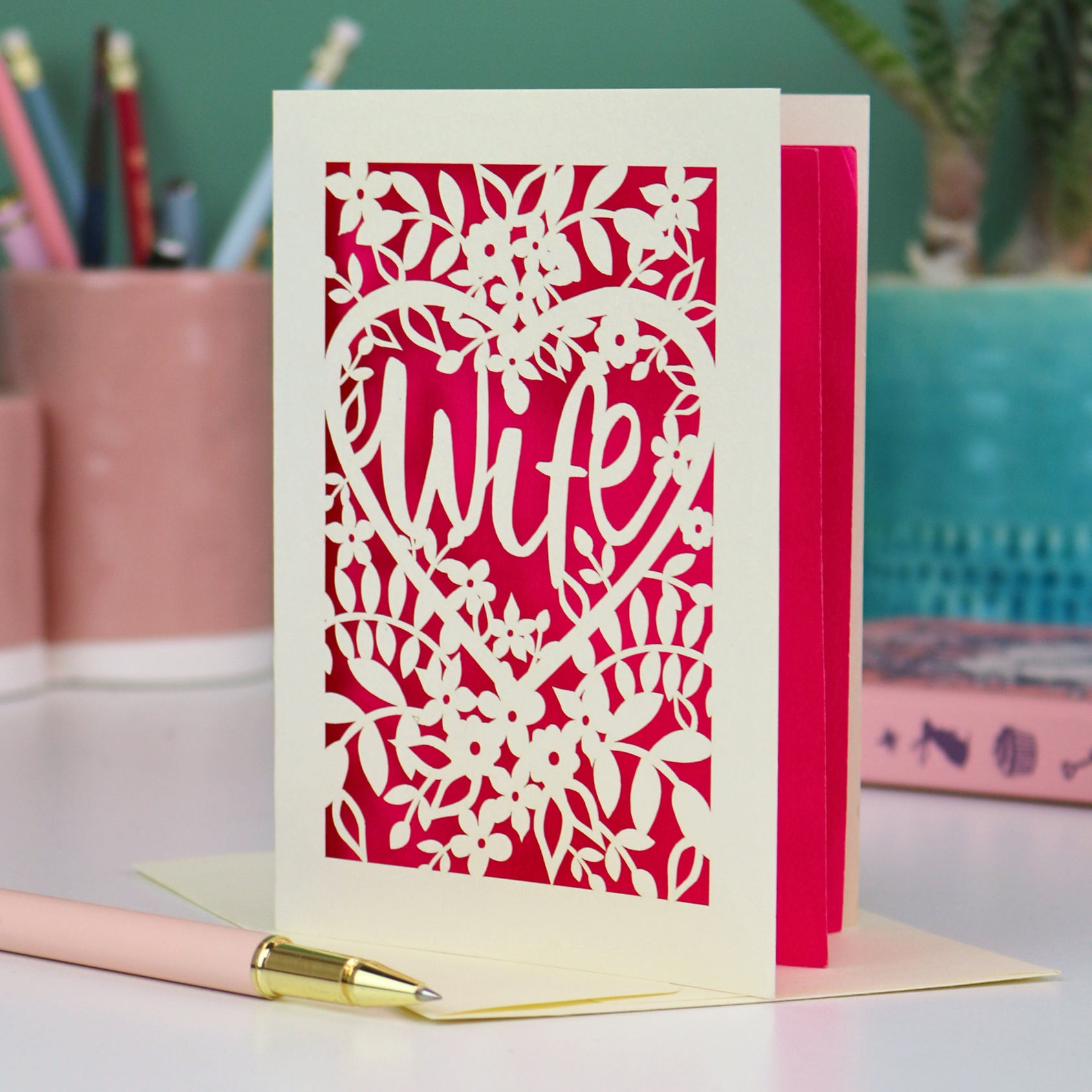Luxury Greeting card with 'wife' in a heart design on a shocking pink background, placed on a desk with stationery items.