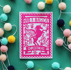 A pink Christmas card with a silhouette of a unicorn and the text 'HAVE A MAGICAL CHRISTMAS' on it, displayed on a green background surrounded by multicolored pom poms.