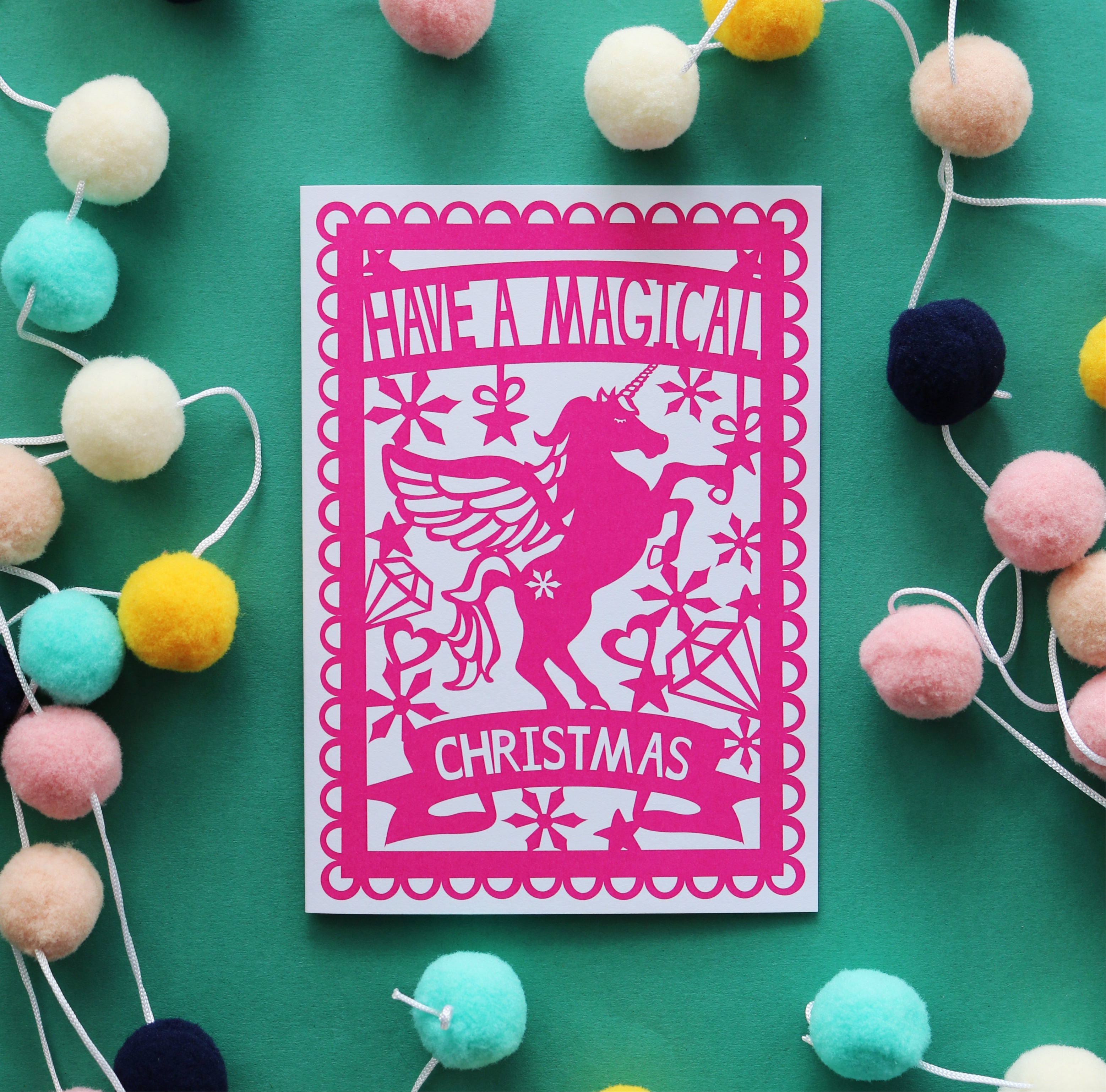 A pink Christmas card with a silhouette of a unicorn and the text 'HAVE A MAGICAL CHRISTMAS' on it, displayed on a green background surrounded by multicolored pom poms.