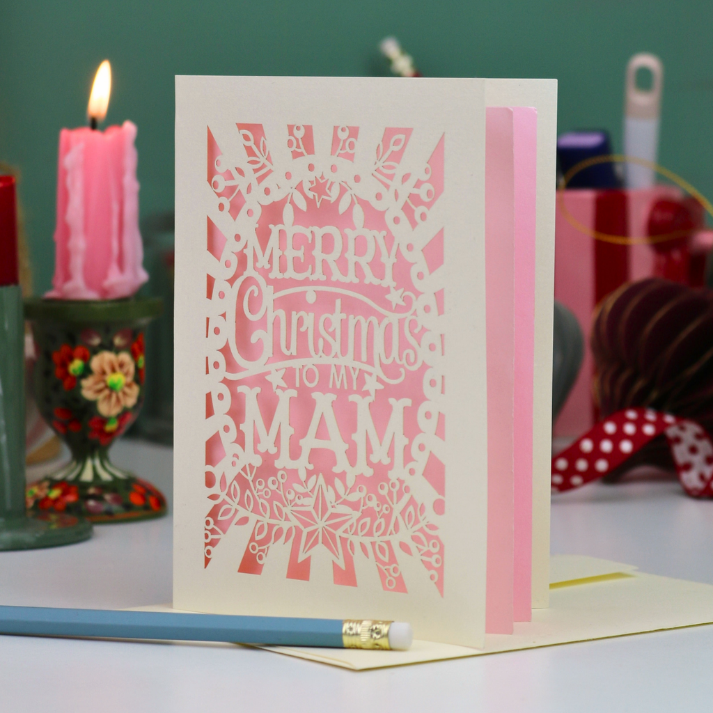 A laser-cut Mam Christmas card with 'Merry Christmas To My Mam' in cut out lettering on a cream cardstock with a candy pink paper backing, standing upright on a table with a red ribbon bookmark, a blue pencil, and a lit pink candle in the background.