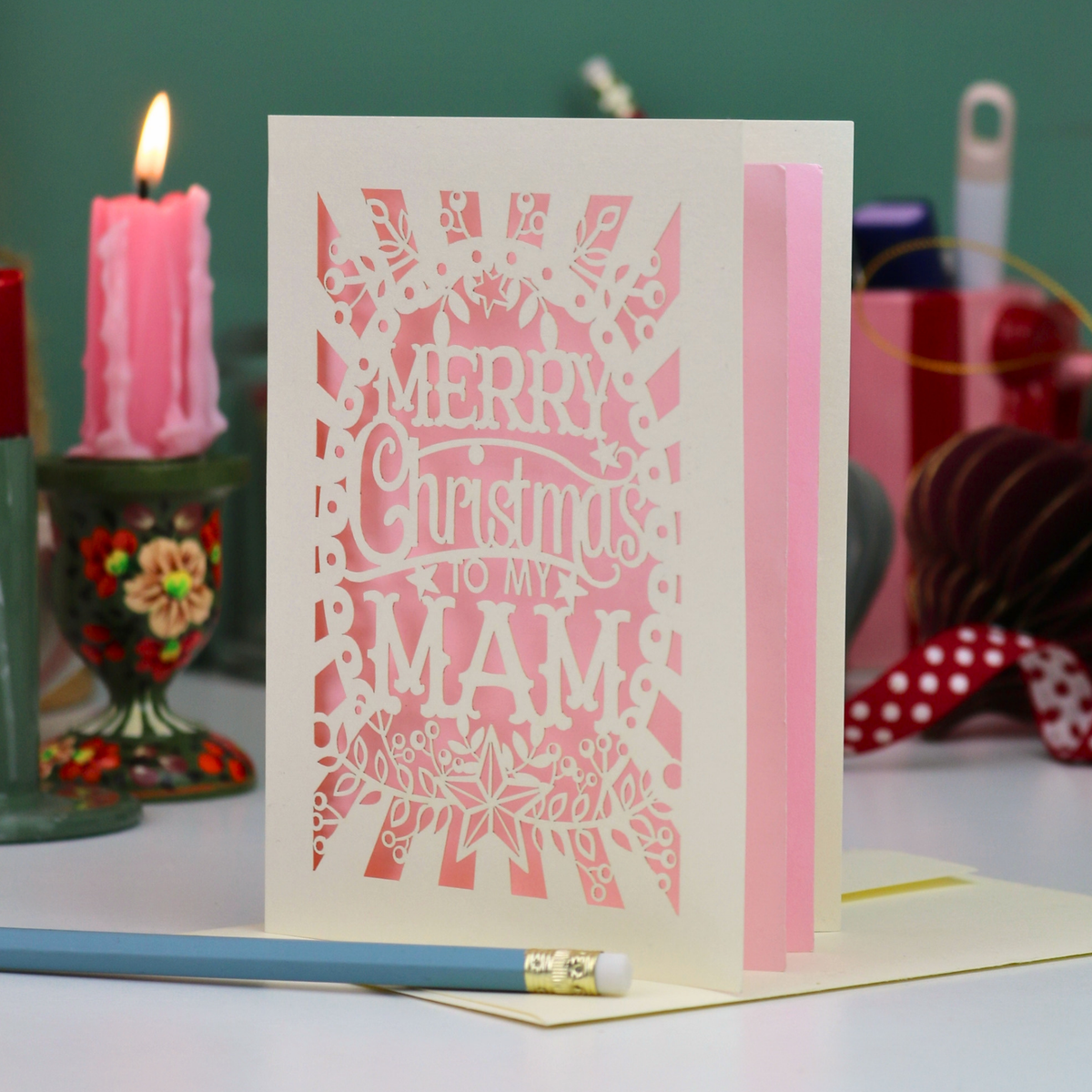 A laser-cut Mam Christmas card with 'Merry Christmas To My Mam' in cut out lettering on a cream cardstock with a candy pink paper backing, standing upright on a table with a red ribbon bookmark, a blue pencil, and a lit pink candle in the background.