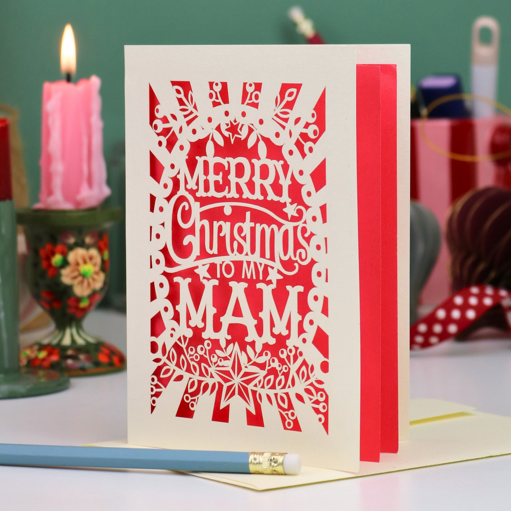 A handmade laser-cut Mam Christmas card with 'Merry Christmas To My Mam' in cut out lettering on a cream cardstock, standing upright on a table with a red ribbon bookmark, a blue pencil, and a lit pink candle in the background.