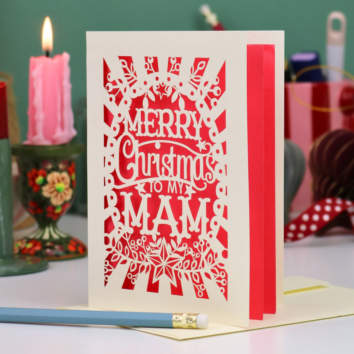A handmade laser-cut Mam Christmas card with 'Merry Christmas To My Mam' in cut out lettering on a cream cardstock, standing upright on a table with a red ribbon bookmark, a blue pencil, and a lit pink candle in the background.