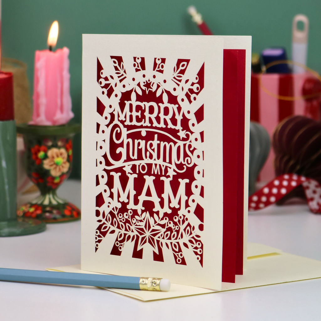 A laser-cut Mam Christmas card with 'Merry Christmas To My Mam' in cut out lettering on a cream cardstock with dark red paper insert, standing upright on a table with a red ribbon bookmark, a blue pencil, and a lit pink candle in the background.