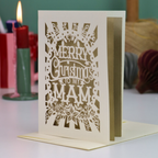 A laser-cut Christmas greeting card with the text 'Merry Christmas to my Mam' and ornate holiday patterns, displayed standing upright on a table with a matching envelope and blurred holiday decorations in the background.