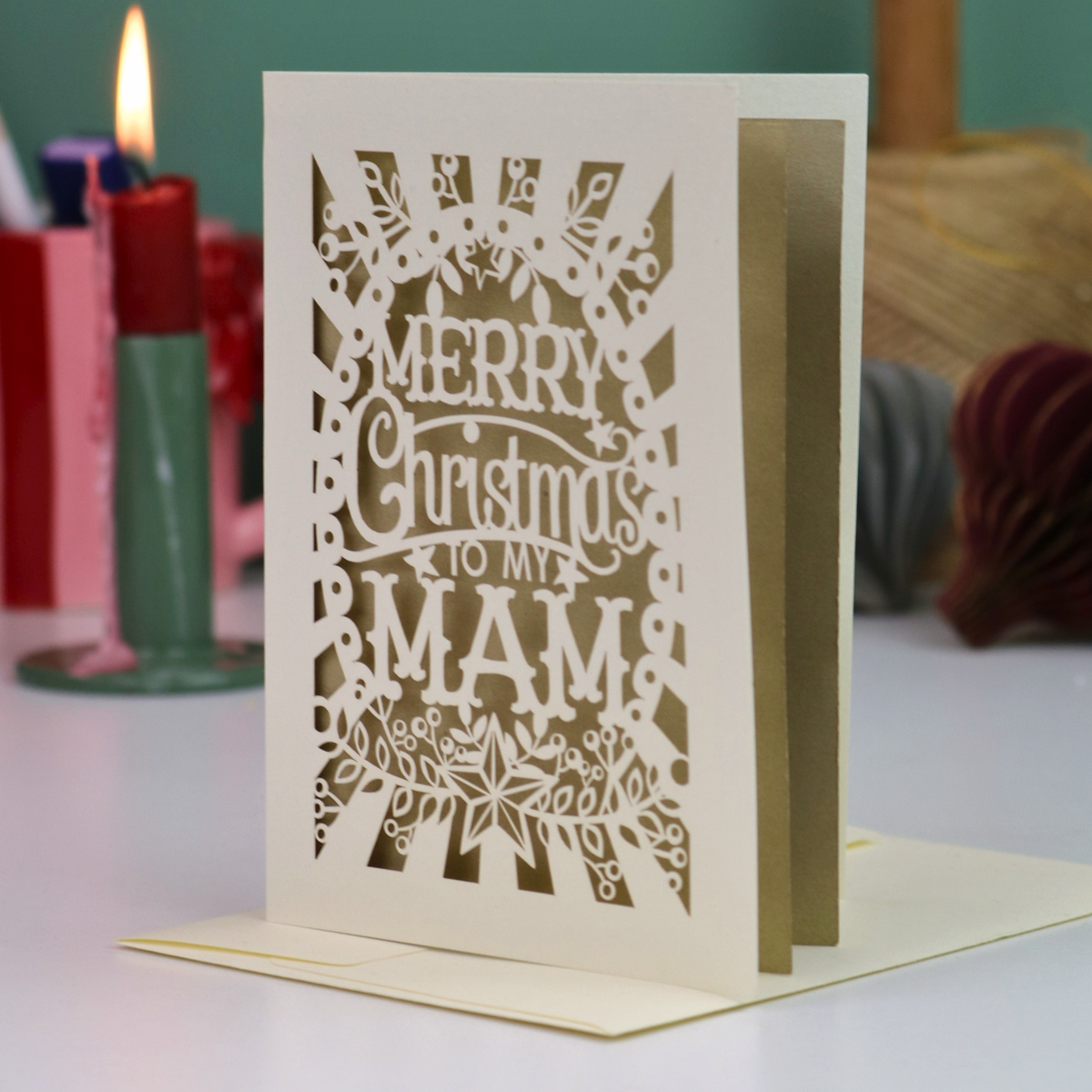 A laser-cut Christmas greeting card with the text 'Merry Christmas to my Mam' and ornate holiday patterns, displayed standing upright on a table with a matching envelope and blurred holiday decorations in the background.