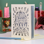 A handmade laser-cut Mam Christmas card with 'Merry Christmas To My Mam' in cut out lettering on a cream cardstock, standing upright on a table with a red ribbon bookmark, a blue pencil, and a lit pink candle in the background.