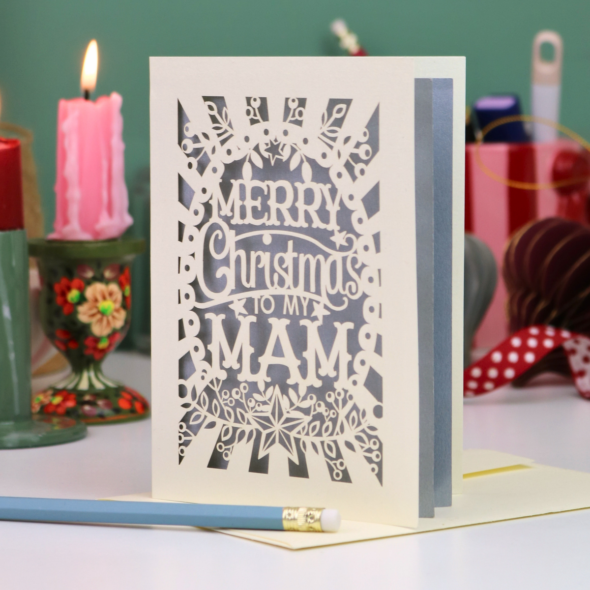 A handmade laser-cut Mam Christmas card with 'Merry Christmas To My Mam' in cut out lettering on a cream cardstock, standing upright on a table with a red ribbon bookmark, a blue pencil, and a lit pink candle in the background.