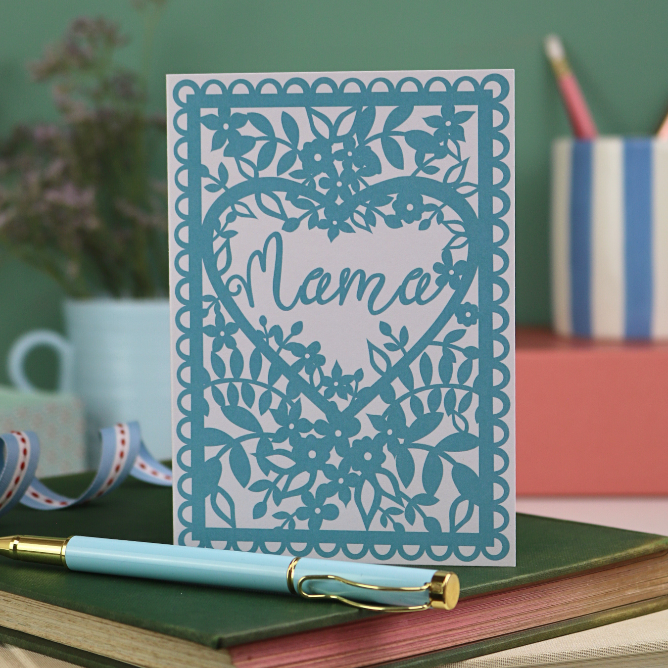 A white greetings card with blue floral design printed and the word "mama" in a script font inside a heart shape. 