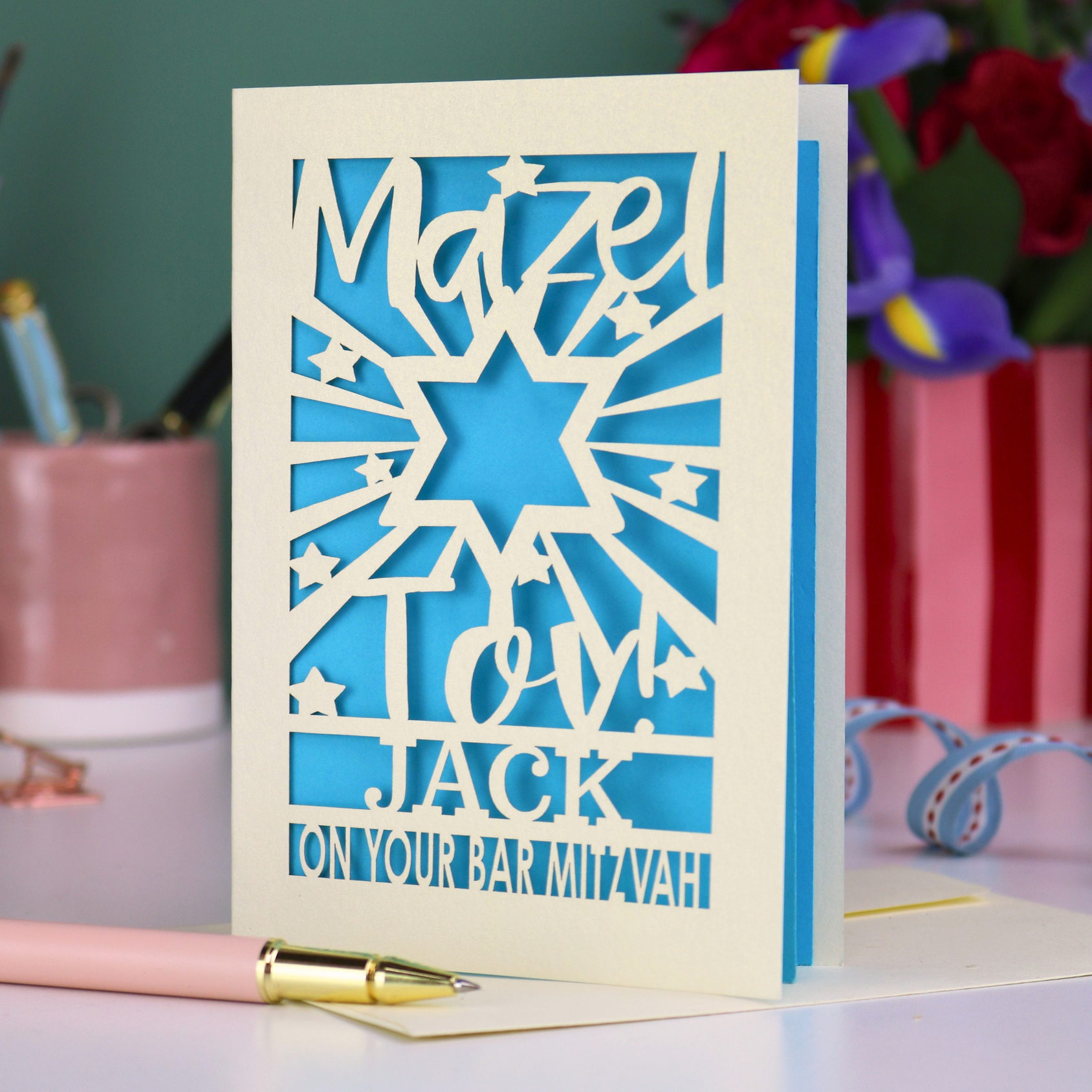 Laser-cut Bar Mitzvah card with blue and cream star design on a desk with flowers and stationery.