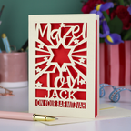 Decorative Laser cut Bar Mitzvah card with cut-out text, 'Mazel Tov Jack on your Bar mitzvah' on a table with stationery items.
